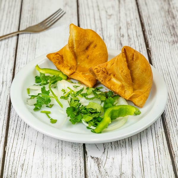 Two golden-brown Tadka Indian Restaurant samosas on a white plate with fresh cilantro, onions, and green peppers.