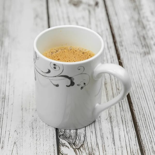 A white mug with a black floral design, filled with frothy Indian chai, sits on a rustic wooden table.