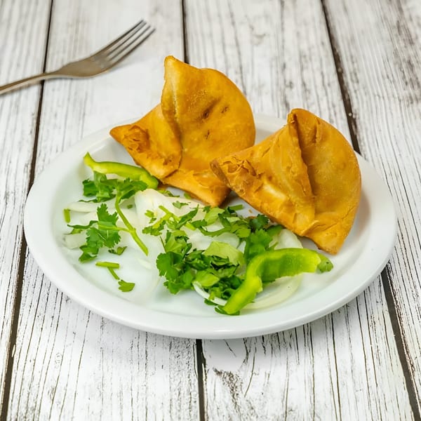 Two golden-brown Tadka Indian Restaurant samosas on a white plate with fresh cilantro, onions, and green peppers.
