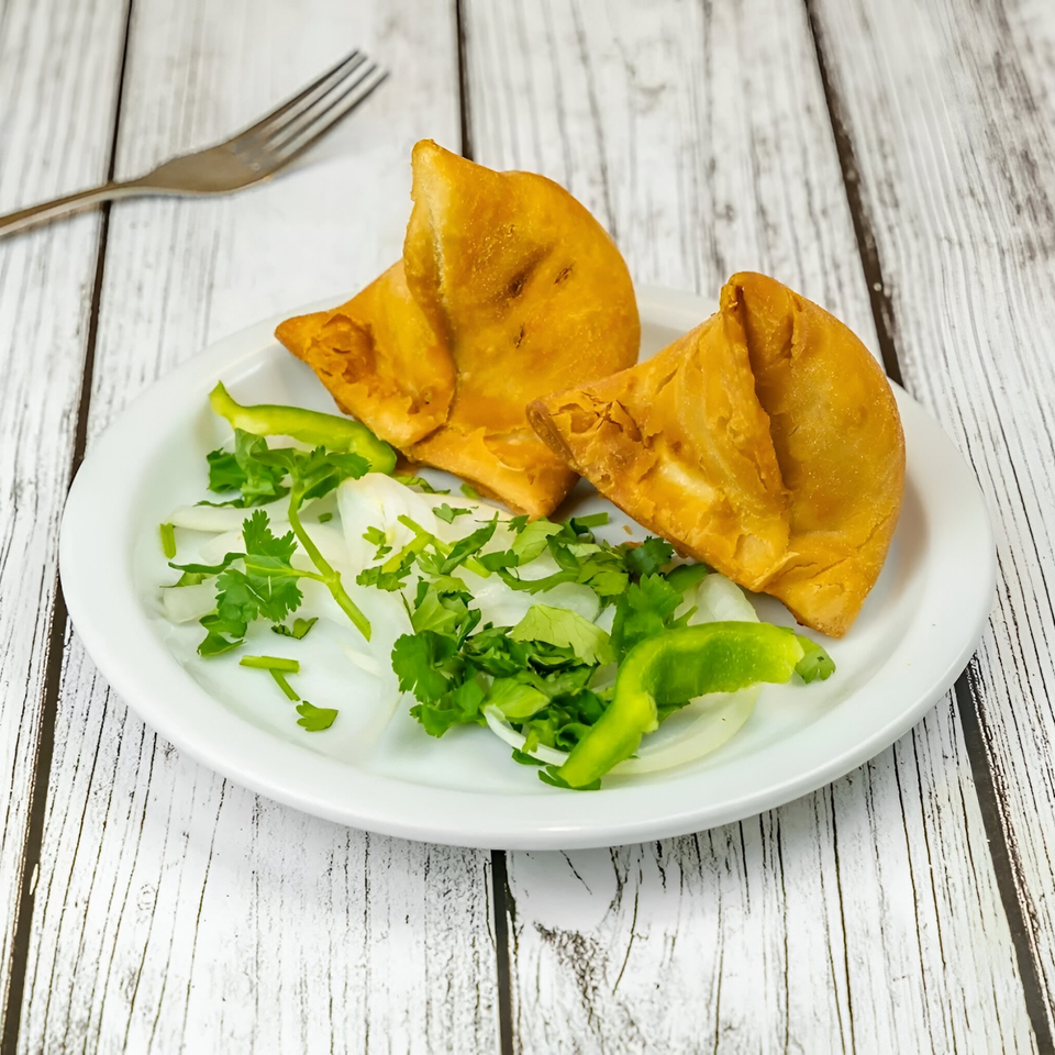 Two golden-brown Tadka Indian Restaurant samosas on a white plate with fresh cilantro, onions, and green peppers.