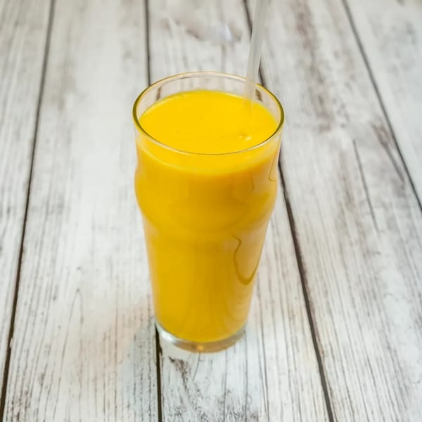 A tall glass of bright yellow mango lassi with a straw on a light wooden table.