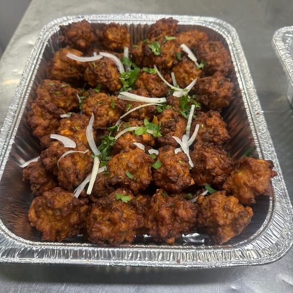 A foil tray filled with golden-brown Indian pakoras, garnished with fresh cilantro and sliced onions.